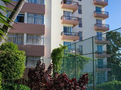 Moderní hotelová budova s balkony obklopená zelenými rostlinami a palmami.