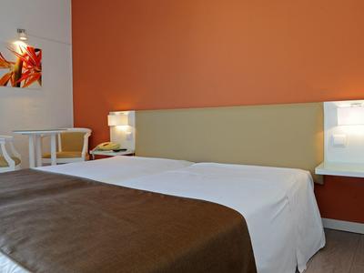 Pohodlný hotelový pokoj s manželskou postelí, hnědou dekou, bílým stolem a oranžovou zdí.