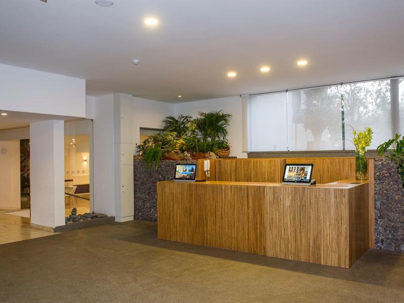 Moderní hotelová recepce s dřevěnou přepážkou a zelenými rostlinami u velkých oken.