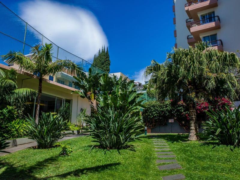 Zelená zahrada s palmami a cestou před hotelovou budovou pod modrou oblohou.