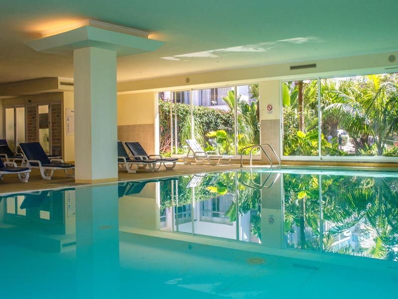 Vnitřní bazén s lehátky a velkými okny s výhledem na tropické rostliny.