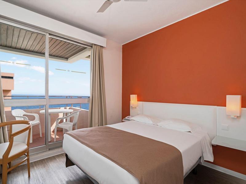 Moderní hotelový pokoj s manželskou postelí, oranžovou zdí, balkonem a výhledem na moře.