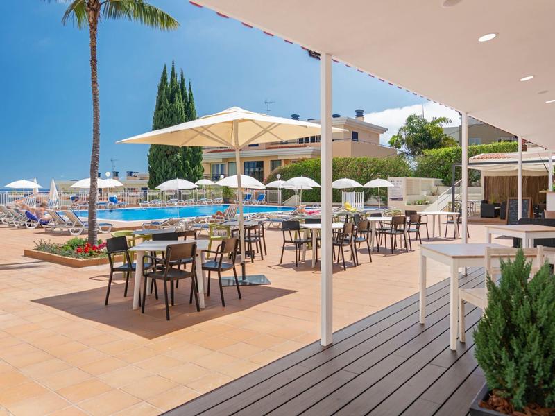 Venkovní prostor hotelu s bazénem, slunečníky a stoly se židlemi na terase.