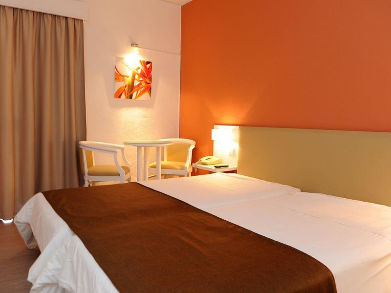 Pohodlný hotelový pokoj s manželskou postelí, oranžovou stěnou a malým stolem se dvěma židlemi.