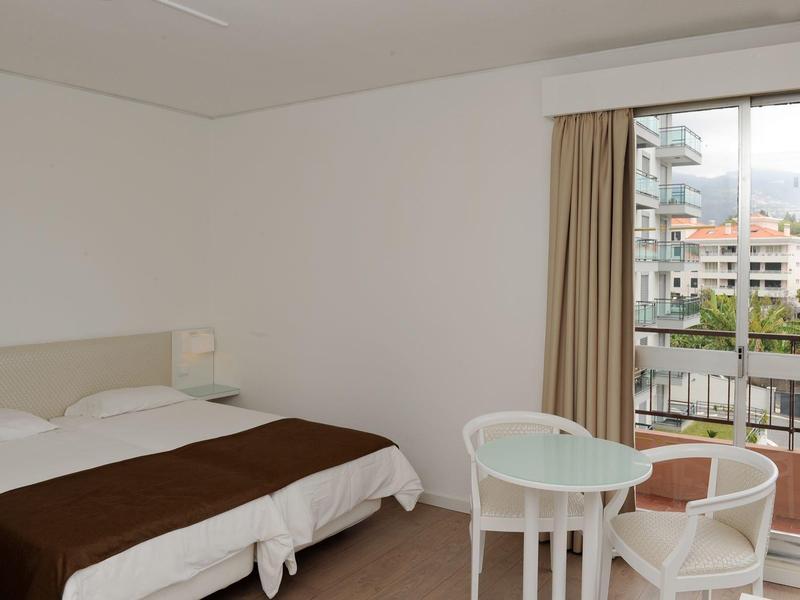 Světý hotelový pokoj s manželskou postelí, malým stolem, dvěma židlemi a balkónem s výhledem.