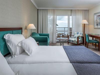 Hotelzimmer mit zwei Betten, Sitzbereich, Schreibtisch und Meerblick durch große Fenster.