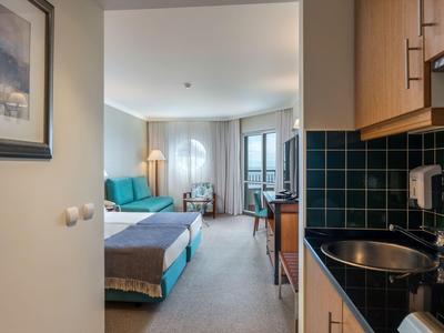 Blick in ein Hotelzimmer mit Küchenzeile, Sofa, Bett und Fenster mit Vorhängen.
