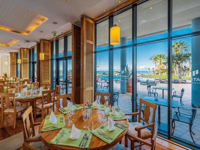 Elegantes Restaurant mit runden Holztischen, grünen Tischdecken und großer Glasfront mit Meerblick.