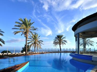 Hotelpool mit Blick auf das Meer und Palmen unter klarem, blauem Himmel.