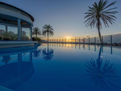 Hotelpool mit Palmen und Glasgeländer bei Sonnenuntergang mit klarer Himmelsspiegelung im Wasser.