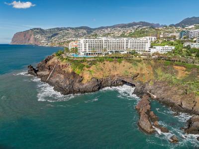 Hotel am Klippenrand mit Blick auf das blaue Meer und bewaldete Hügel im Hintergrund.