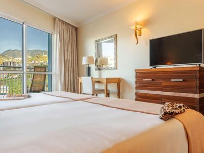 Hell eingerichtetes Hotelzimmer mit Doppelbett, Schreibtisch, Fernseher und Balkon mit Bergblick.
