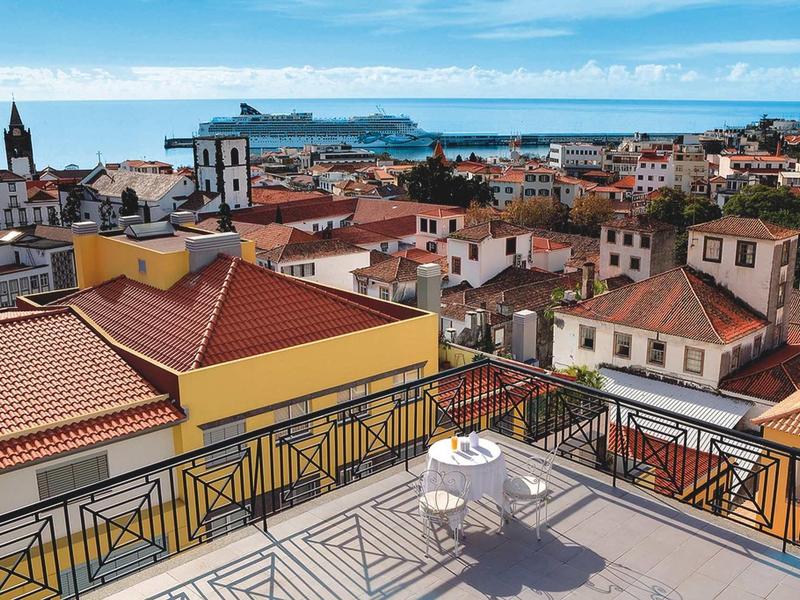 Vista da una terrazza con tavolo e sedie su una città mediterranea e il mare.