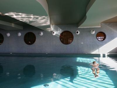 Helles Hallenbad mit Fenstern, blauer Wasseroberfläche und Person, die im Pool schwimmt.