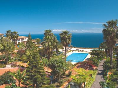 Blick auf einen Pool umgeben von Palmen und Grünfläche mit Meer und blauem Himmel im Hintergrund.