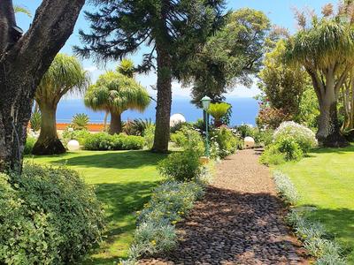 Ein Gartenweg aus Steinen führt durch grüne Wiesen zu Bäumen und blauem Meer im Hintergrund.
