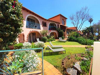 Großes zweistöckiges Haus mit roten Ziegeln, grüner Garten mit Liegestühlen und bunten Blumen.