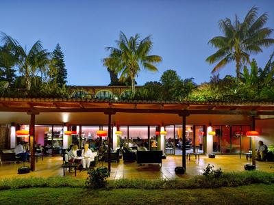 Offenes Restaurant mit Holzdach, vielen Gästen, grüner Garten- und Palmenkulisse bei Abenddämmerung.