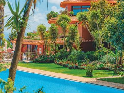 Hotel z niebieskim basenem otoczonym palmami i zielonym ogrodem pod czystym niebem.