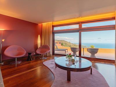 Nowoczesny salon z dużymi oknami i widokiem na morze, dwa różowe fotele i drewniana podłoga.