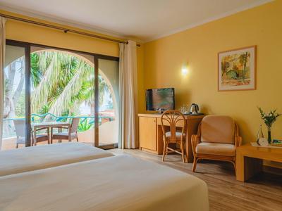 Pokój hotelowy z podwójnym łóżkiem, biurkiem, telewizorem i balkonem z widokiem na palmy.