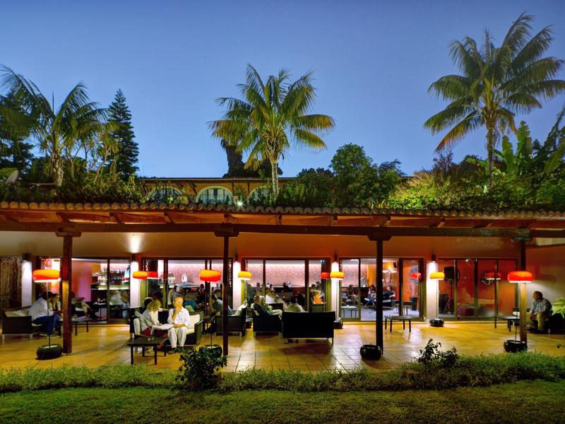 Otwarta restauracja z stołami i krzesłami, oświetlona o zachodzie słońca, otoczona palmami.