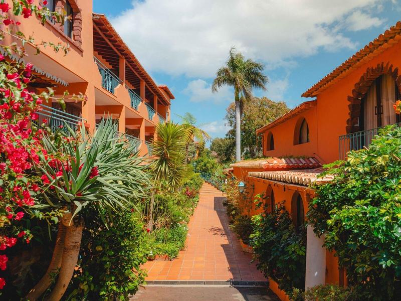 Ścieżka między pomarańczowymi budynkami z bujną roślinnością i palmami pod niebieskim niebem.