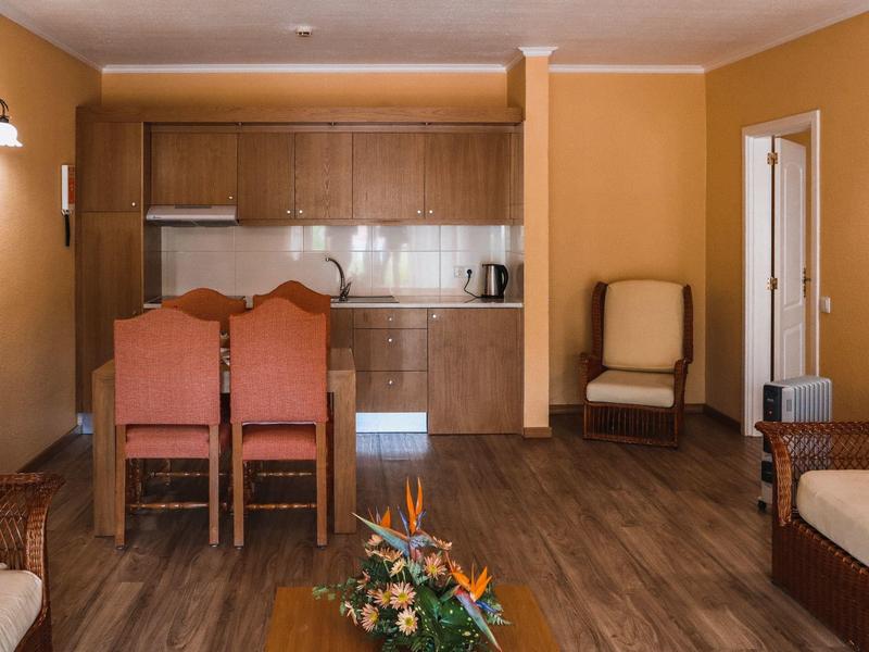 Jasnobrązowy salon z aneksem kuchennym, dwoma krzesłami, fotelem i drewnianą podłogą.