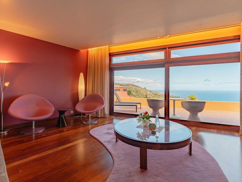 Nowoczesny salon z dużymi oknami i widokiem na morze, dwa różowe fotele i drewniana podłoga.