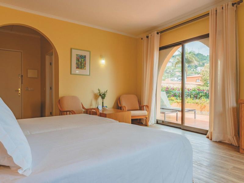 Jasny pokój hotelowy z podwójnym łóżkiem, miejscem do siedzenia i dużym oknem z widokiem na taras.