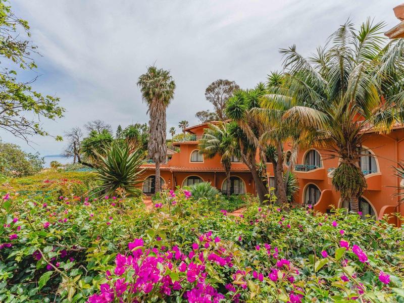 Widok na hotel z pomarańczową elewacją otoczony palmami i kolorowymi kwiatami.