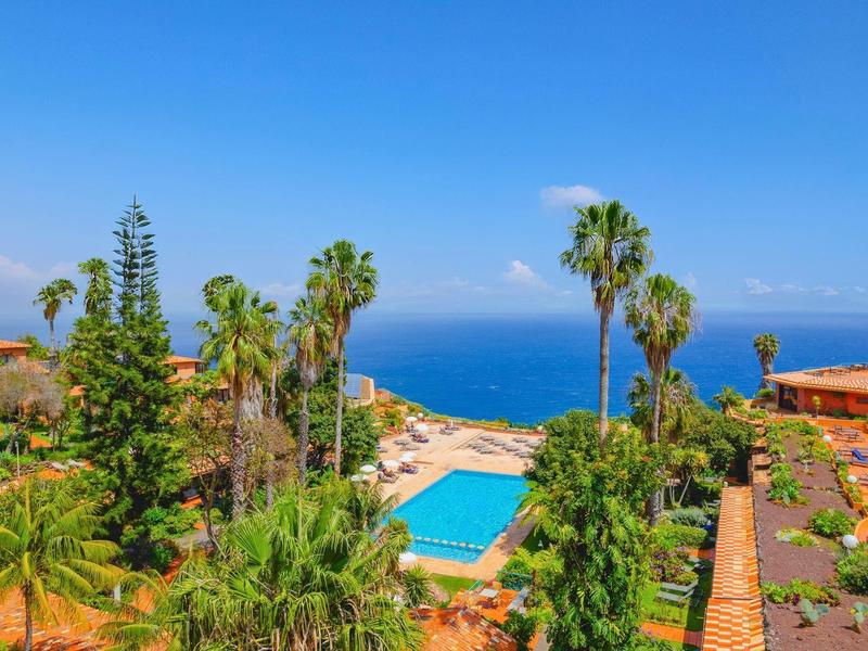 Widok na hotelowy basen z palmami i morzem w tle w słoneczny dzień.