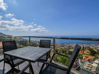 Balcon avec table et quatre chaises donnant sur la mer et une ville sous un ciel bleu.