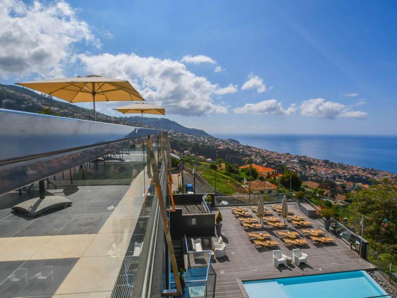 Vue depuis une terrasse d'hôtel avec parasols sur une piscine et la côte par temps ensoleillé.