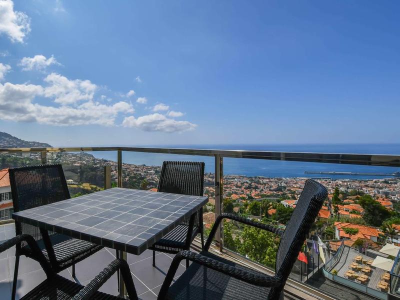 Balcon avec table et quatre chaises donnant sur la mer et une ville sous un ciel bleu.