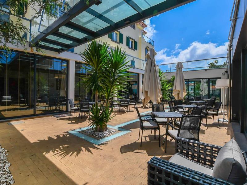 Terrasse d'hôtel ensoleillée avec des chaises en métal et des plantes vertes sous un ciel bleu.