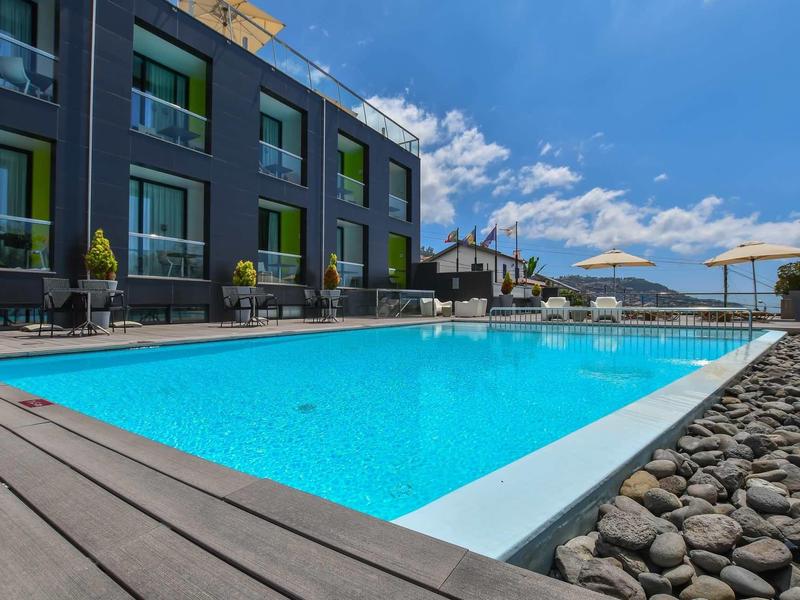 Espace piscine moderne avec transats, parasols et bâtiment d'hôtel adjacent sous un ciel bleu.