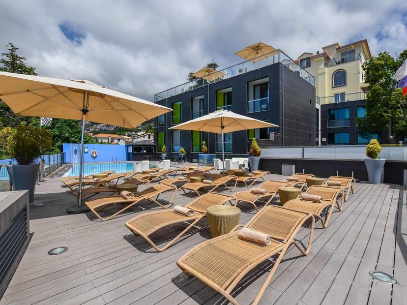 Terrasse ensoleillée avec chaises longues et parasols devant des bâtiments d'hôtel modernes sous un ciel couvert.