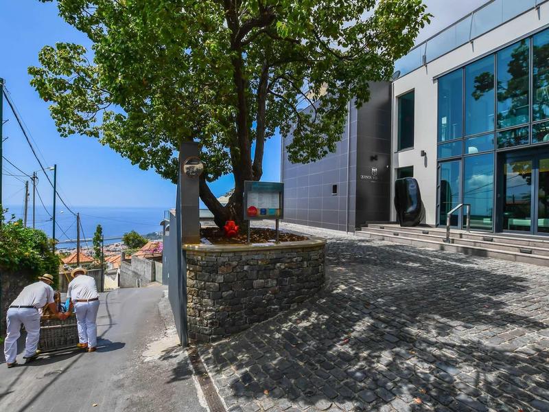 Deux personnes descendent une rue raide à côté d'un bâtiment moderne avec façade en verre et vue sur la mer.