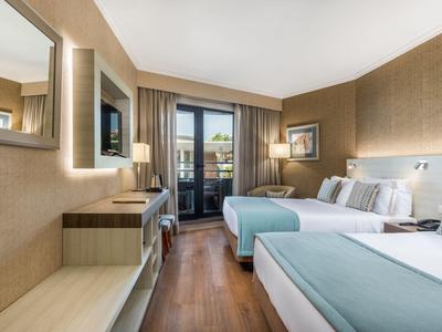 Hotel room with two double beds, wooden floor, and balcony overlooking buildings.