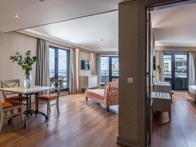 Modern furnished hotel rooms with wooden floors, table, chairs, and large windows overlooking the city.