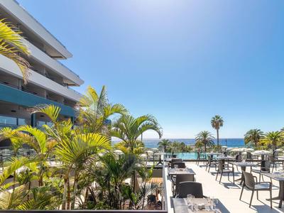 Hotel terrace with tables, chairs, palm trees, and sea view under a clear sky.
