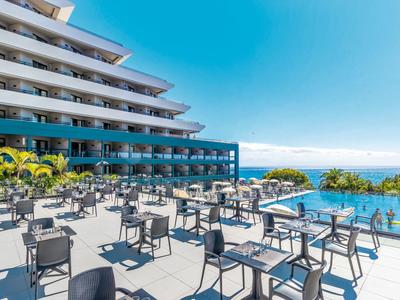 Modern hotel terrace with tables and chairs, view of pool and sea under clear sky.