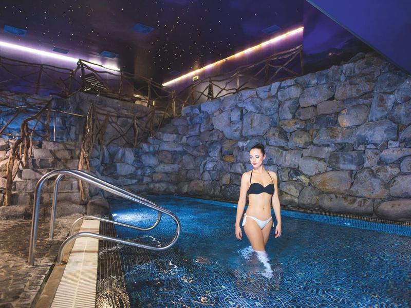 Woman in black bikini steps into a lit hotel pool with stone wall background.