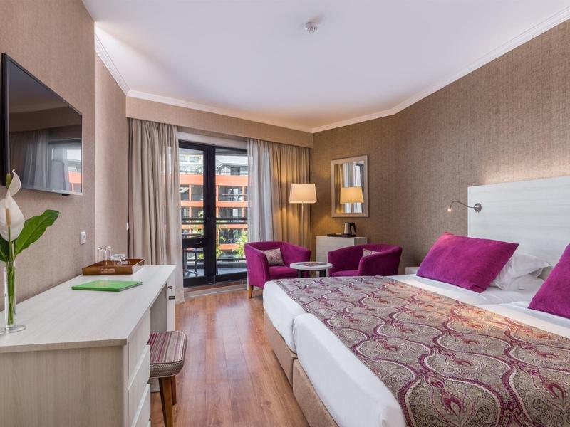 Modern hotel room with bed, seating area, TV, and balcony overlooking buildings.