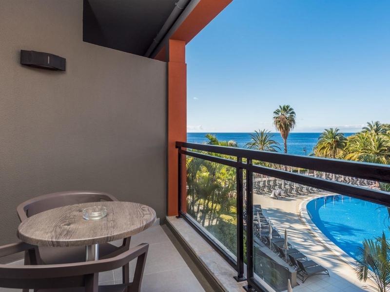 Balcony with table and chairs overlooking pool, palm trees, and the sea at the hotel