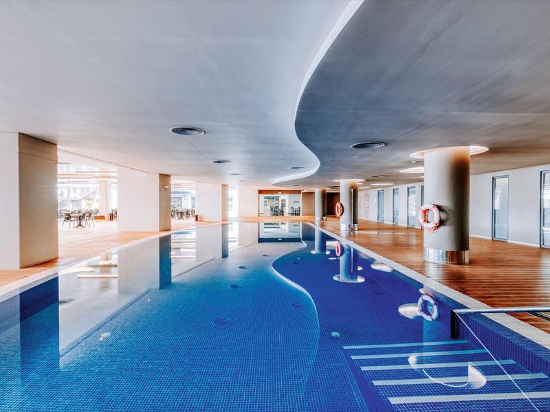 Modern indoor pool with curved ceiling and seating areas in bright wellness space.