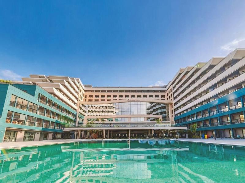 Modern hotel with large outdoor pool and multi-story balconies under clear sky.