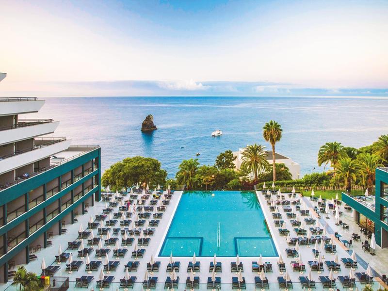 Large hotel pool with many lounge chairs and a view of the sea and a rock in the water.