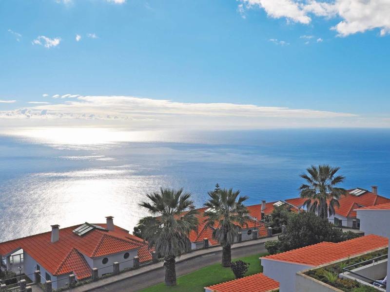 Vista di un complesso costiero con tetti rossi, palme e mare calmo sotto un cielo blu.